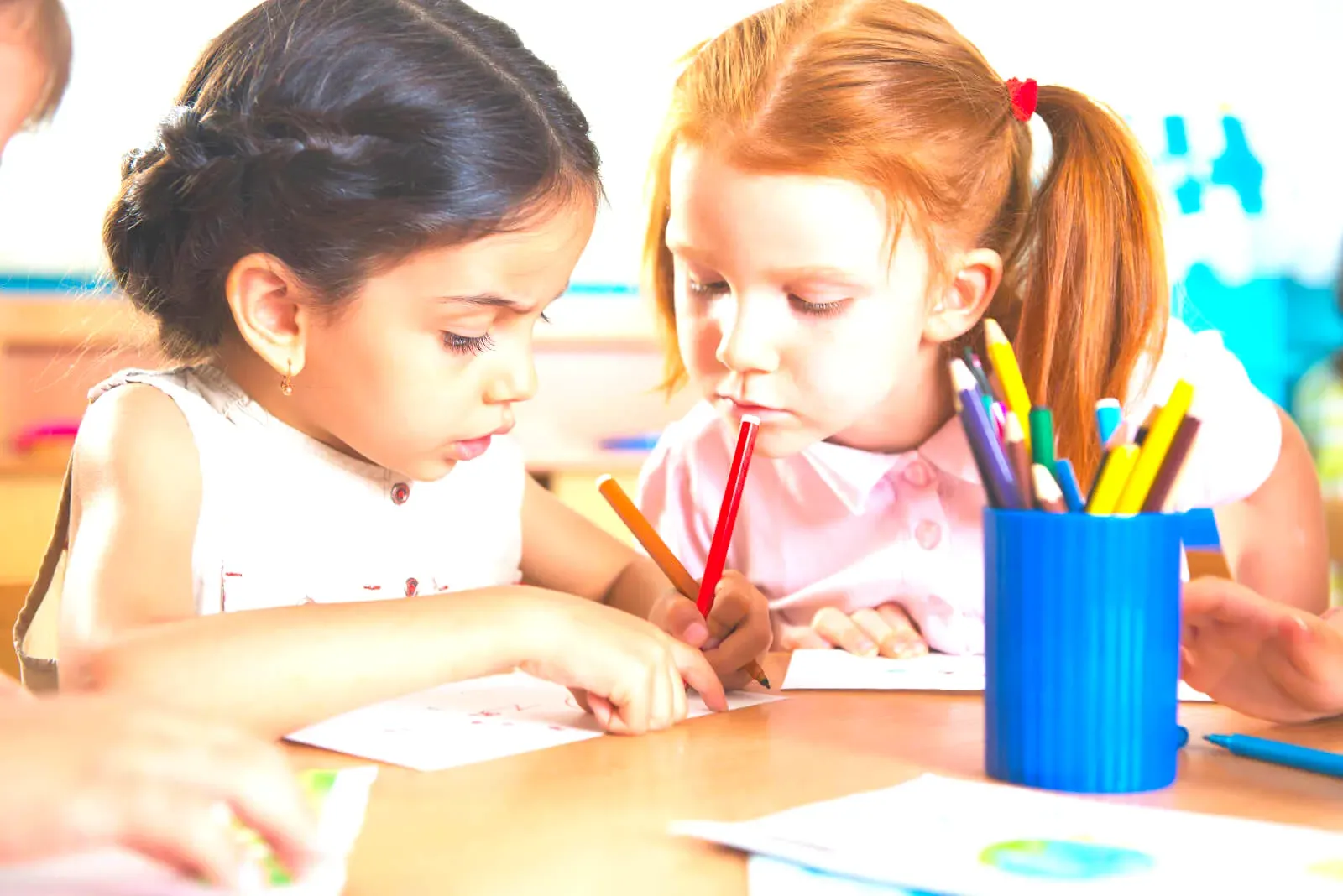 Two girls concentrating on a drawing at a table, with a pot of coloured pencils